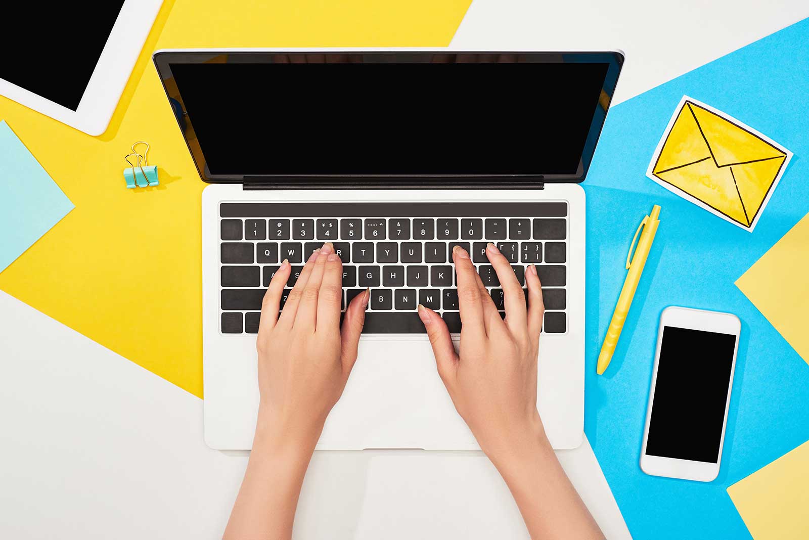 Overhead view of female typing on laptop surrounded by blue and yellow papers.